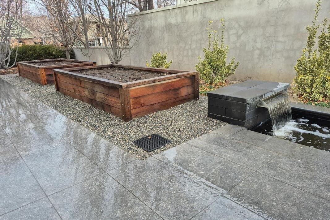 Two redwood raised beds on pea gravel beside a dark stone water feature and a wet stone patio